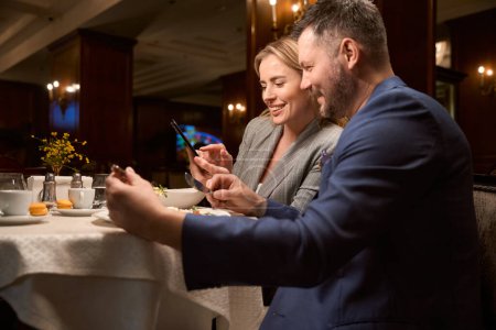 Téléchargez les photos : Joyeux couple marié qui dîne au lobby-bar et regarde de nouvelles photos de mariage sur smartphone, ayant des souvenirs amusants et agréables - en image libre de droit