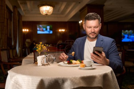 Foto de Hombre de negocios guapo comiendo sabroso almuerzo y beber café leyendo mensaje importante en el teléfono inteligente - Imagen libre de derechos