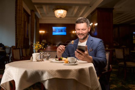 Foto de Hombre feliz sonriendo, charlando en el teléfono inteligente en citas aplicación y almorzando en el lobby-bar - Imagen libre de derechos