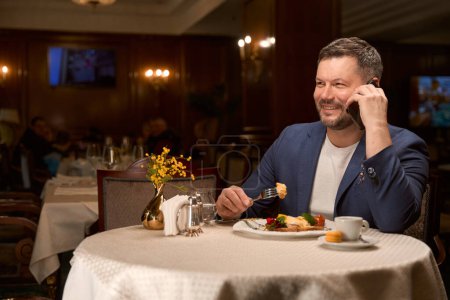 Photo for Lonely handsome man eating delicious meal at lobby-bar and smiling, talking on smartphone, flirt - Royalty Free Image