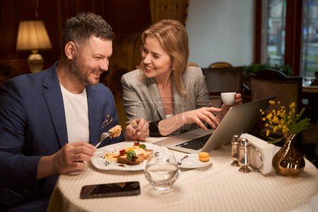 Photo for Cheerful spouses smiling looking at photos from vacation on laptop during dinner at restaurant - Royalty Free Image