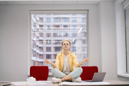 Photo for Young woman office worker meditating sitting in lotus asana on work desk, relaxing and opening her mind for ideas, preparing for new case - Royalty Free Image