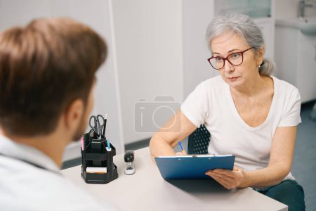 Foto de Paciente de edad avanzada llena un formulario médico en una cita con un médico, un terapeuta dirige una cita en un centro médico - Imagen libre de derechos
