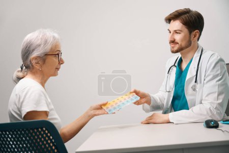 Foto de Trabajador médico da una caja de pastillas con pastillas a un paciente de edad avanzada, el médico tiene un estetoscopio en el cuello - Imagen libre de derechos