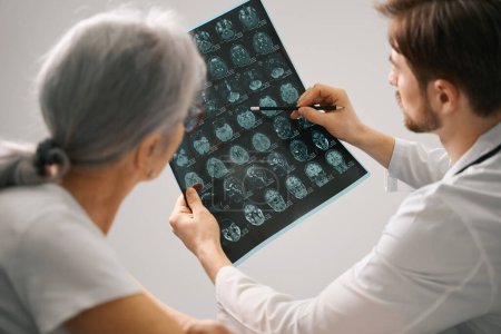 Téléchargez les photos : Docteur diagnosticien tient un scanner IRM du cerveau dans ses mains, il consulte une dame âgée - en image libre de droit
