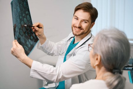Foto de Agradable joven doctor está sosteniendo una resonancia magnética en sus manos, él está consultando a una anciana - Imagen libre de derechos