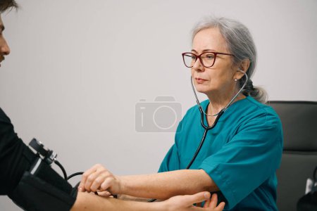Photo for Woman practitioner measures blood pressure to young bearded man, doctor uses tonometer and stethoscope - Royalty Free Image