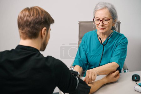 Photo for Attentive female therapist measures blood pressure to a young patient, man in casual clothes - Royalty Free Image