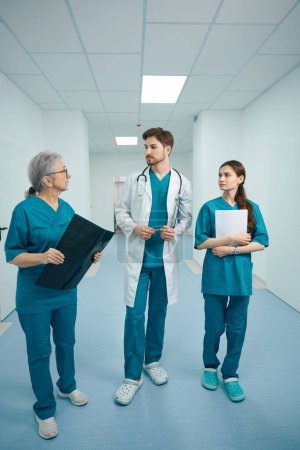 Photo for Three colleagues of doctors are walking along the hospital corridor, the doctors have folders in their hands, MRI scans, documents - Royalty Free Image