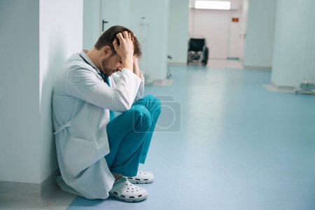 Upset young intern squatting in hospital corridor with wheelchair in the background