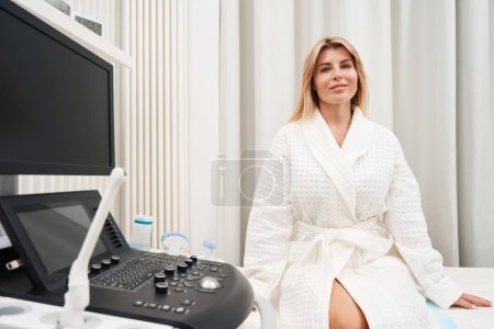 Photo for Smiling female patient in a bathrobe sits on a couch near ultrasound machine, woman on consultation in the diagnostic department - Royalty Free Image