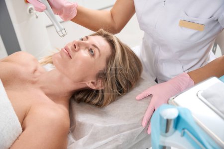Charming lady on laser skin resurfacing procedure, female beautician in medical uniform uses CO2 laser