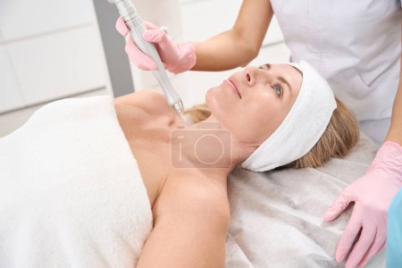 Photo for Patient is located on a cosmetology couch for laser skin resurfacing procedure, female in medical uniform uses a modern device - Royalty Free Image