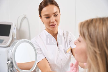 Foto de El médico cosmetólogo examina al cliente de la clínica de medicina estética, la mujer se mira en el espejo - Imagen libre de derechos
