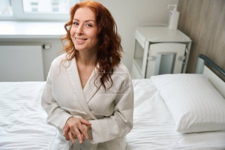 Foto de Sonriente paciente femenina frente a la cámara mientras está sentado en la cama enferma en la habitación moderna del hospital - Imagen libre de derechos