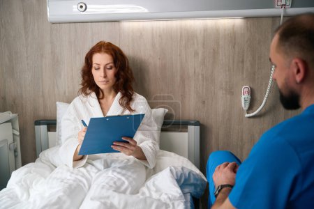 Téléchargez les photos : Femme mûre cliente allongée sur un lit de malade confortable et signant des documents dans la chambre du centre de santé - en image libre de droit