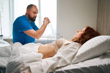 Téléchargez les photos : Rousse visiteuse en train d'être examinée par un médecin alors qu'elle était allongée sur un lit confortable dans un centre de santé moderne - en image libre de droit