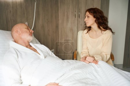 Foto de Amable esposa con el pelo rojo visitando a su apuesto marido en la sala de rehabilitación del hospital - Imagen libre de derechos