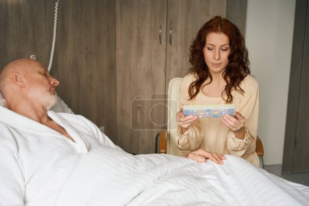 Foto de Mujer bonita sentada en una silla moderna y sosteniendo la caja de pastillas mientras su marido pensativo yacía en la cama en el interior - Imagen libre de derechos
