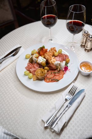 Foto de Plato con queso, uvas, jamón y otros aperitivos servido con copas de vino tinto en la mesa blanca en el restaurante - Imagen libre de derechos