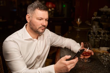 Foto de Hombre barbudo guapo usando teléfono inteligente mientras está sentado solo con un vaso de whisky en el mostrador de bar - Imagen libre de derechos