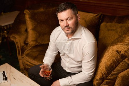 Téléchargez les photos : Bel homme sérieux se détendre avec un verre d'alcool cher tout en étant assis sur le canapé dans le restaurant de luxe - en image libre de droit