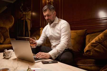 Téléchargez les photos : Bel homme barbu travaillant sur ordinateur portable tout en étant assis seul avec un verre de whisky sur le canapé dans le restaurant - en image libre de droit