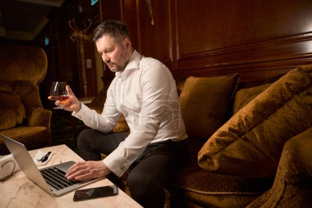 Téléchargez les photos : Homme d'affaires confiant naviguant quelque chose sur ordinateur portable tout en ayant le temps seul avec un verre de brandy sur le canapé dans le restaurant - en image libre de droit