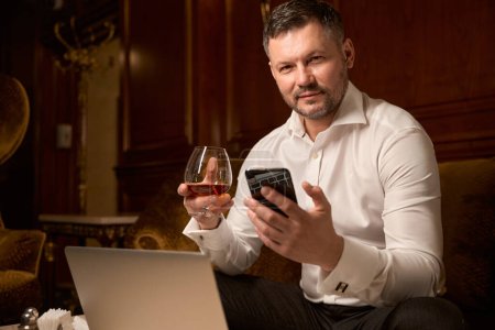 Téléchargez les photos : Homme sérieux confiant textos avec sa femme sur smartphone tout en ayant du bon temps seul avec un verre de brandy sur le canapé dans le restaurant - en image libre de droit