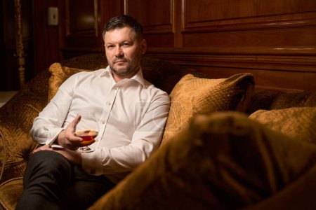 Téléchargez les photos : Élégant invité masculin buvant brandy cher tout en passant du temps seul sur le canapé de luxe dans le restaurant - en image libre de droit