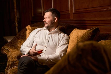 Téléchargez les photos : Beau invité masculin assis avec un verre de cognac cher sur le canapé de luxe dans le restaurant - en image libre de droit
