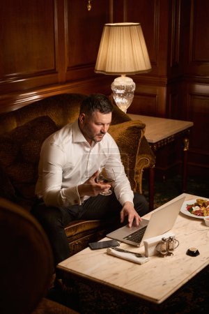 Téléchargez les photos : Homme d'affaires sérieux tapant sur ordinateur portable tout en étant assis par lui-même avec du cognac sur le canapé de luxe dans le restaurant - en image libre de droit