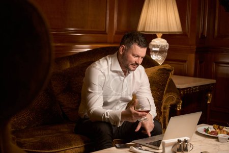 Téléchargez les photos : Homme d'affaires adulte bavardant avec des partenaires sur un ordinateur portable tout en tenant un verre de cognac sur le canapé dans le restaurant de luxe - en image libre de droit