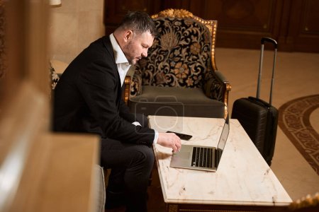 Téléchargez les photos : Beau-homme relaxant sur un fauteuil et naviguant quelque chose sur un ordinateur portable à la table près des bagages dans le hall de l'hôtel - en image libre de droit