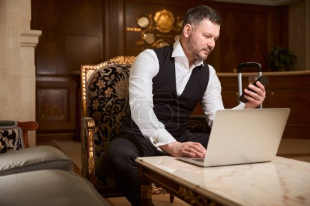 Téléchargez les photos : Homme confiant en costume noir se reposant tout en textant au téléphone près de l'ordinateur portable sur la table dans le hall de l'hôtel - en image libre de droit