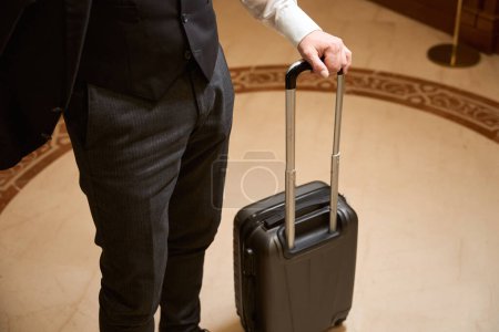 Foto de Foto recortada del hombre de pie con la maleta negra en el vestíbulo del hotel - Imagen libre de derechos
