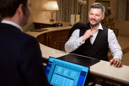 Foto de Guapo hombre de negocios elegante hablando con la recepcionista masculina mientras se registra en el hotel - Imagen libre de derechos