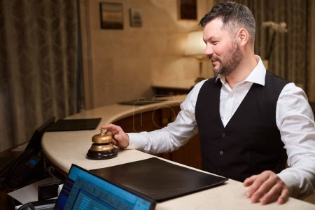 Téléchargez les photos : Un bel homme souriant sonne la cloche du service doré à la réception blanche de l'hôtel de luxe - en image libre de droit