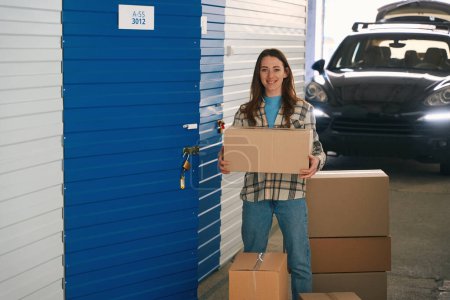 Photo for Smiling lady is holding a large box near open door of the pantry in the storage warehouse. In the background is standing a car with open trunk - Royalty Free Image