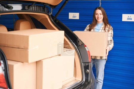 Téléchargez les photos : Jeune femme se tient près du coffre ouvert d'une voiture avec des boîtes dans l'entrepôt et tenant une grande boîte en carton - en image libre de droit