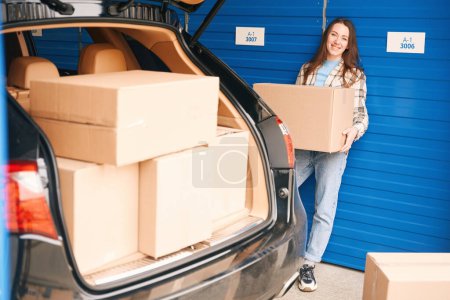 Téléchargez les photos : Jeune femme se tient près du coffre ouvert d'une voiture avec des boîtes dans l'entrepôt et tenant une grande boîte en carton - en image libre de droit