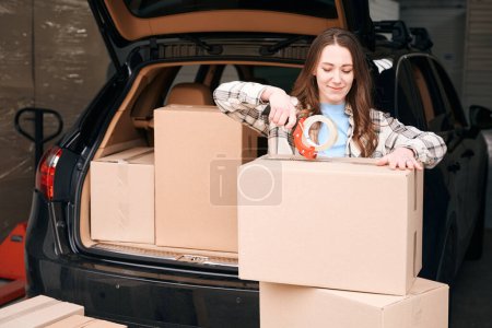 Photo for Cute lady is packing cardboard boxes into the trunk of car in a storage warehouse - Royalty Free Image