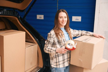 Photo for Smiling lady is packing cardboard boxes into the trunk of car in a storage warehouse - Royalty Free Image