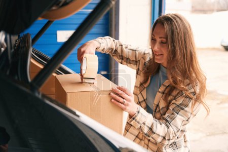 Photo for Smiling lady is packing cardboard boxes with scotch into the trunk of car in the warehouse - Royalty Free Image