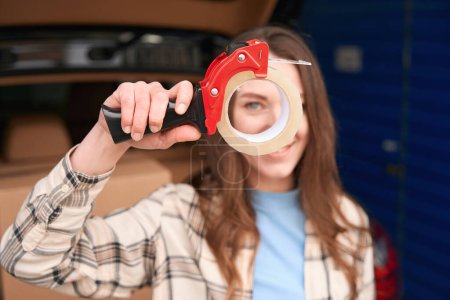 Photo for Smiling lady is holding tape gun with scotch and standing near the open trunk of a car with boxes into it - Royalty Free Image
