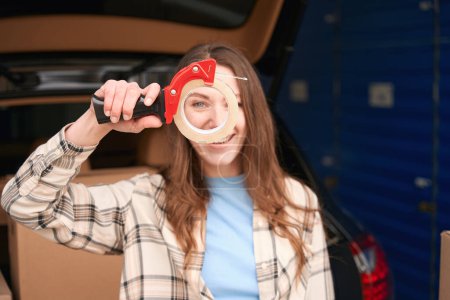 Téléchargez les photos : Femme souriante tient dans le pistolet à ruban à main avec du scotch et debout près du coffre ouvert d'une voiture avec des boîtes dedans - en image libre de droit
