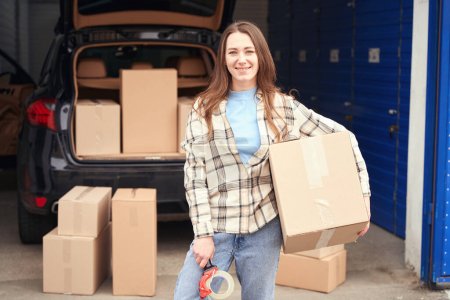Smiling lady is standing near the open trunk of a car with boxes into warehouse. She is holding a big cardboard box and tape gun