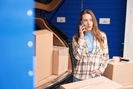 Photo for Young woman is calling with mobile and standing near the trunk of car in a storage warehouse. Cardboard boxes with things for moving are in the trunk and next to the car - Royalty Free Image
