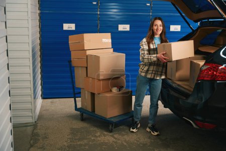 Photo for Smiling woman is puting cardboard boxes in the trunk of a car into warehouse with self storage unit - Royalty Free Image