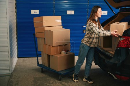 Photo for Young woman is puting cardboard boxes in the trunk of a car from platform truck in storage warehouse - Royalty Free Image